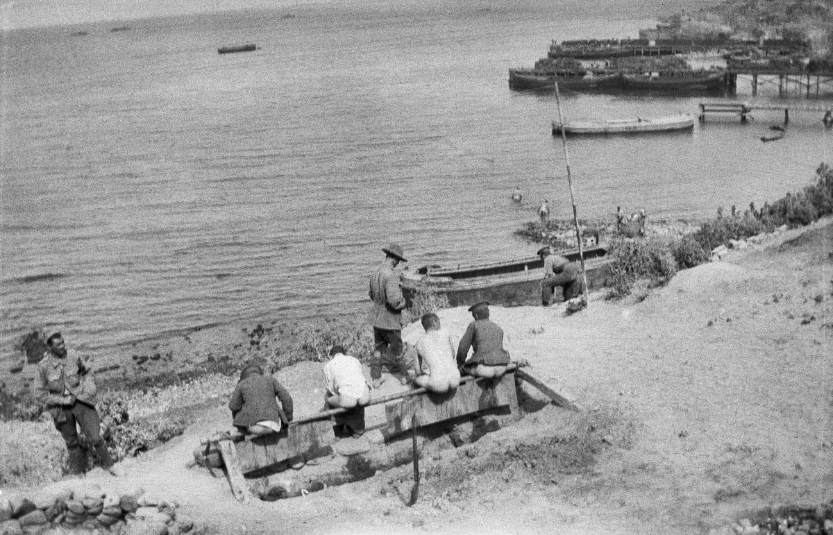Four unidentified men using a latrine high above the beach at Anzac Cove. Obviously dug to provide the users with the safest, best, and most practicable view, it faces the sea.